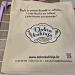 Bež platnena vrećica s tiskom logotipa Dobra kuhinja i ilustracijama povrća (mrkve i luka). Sadrži poruku o zdravoj prehrani i kontakt informacije, uključujući web stranicu www.dobrakuhinja.hr i QR kod. Praktična i ekološka vrećica za promociju.