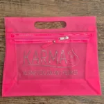 Ružičasta plastična torbica s prozirnim dijelovima i patentnim zatvaračem, na kojoj je srebrni natpis 'KARMA' te dodan opis 'Kozmetički salon i pediker', uz minimalistički grafički prikaz ženskog profila. Dizajn torbice namijenjen je za promociju i praktičnu upotrebu u kozmetičkoj industriji