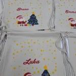 Slika prikazuje personalizirane platnene vrećice s božićnim motivima, uključujući Djeda Božićnjaka, božićno drvce i zvjezdice. Na svakoj vrećici ispisano je ime, poput Aria, Bruno, Emma, Luka i Rino, čime se naglašava personalizacija i prikladnost za darivanje u blagdansko vrijeme.